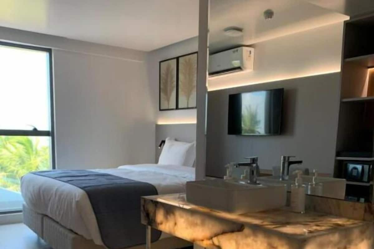 A modern hotel room with a bed, a large window, a TV mounted on the wall, and a bathroom with a vanity and sink.