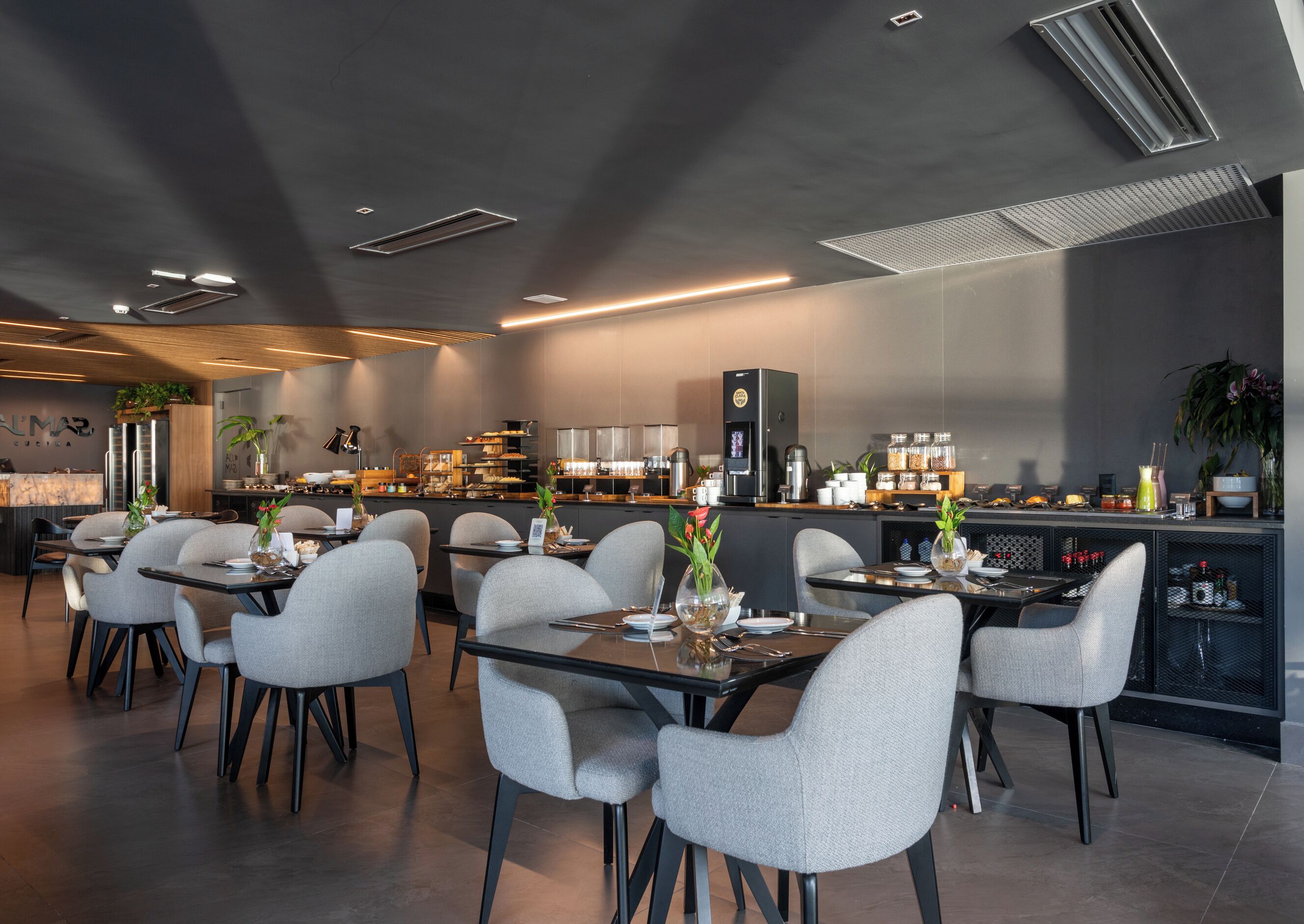 A modern restaurant with a dark interior, grey chairs, and a bar area with various items.