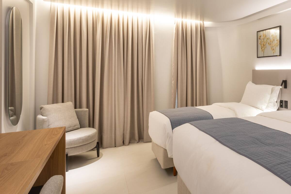 A modern hotel room with a large bed, a chair, a desk, and a large window with curtains.
