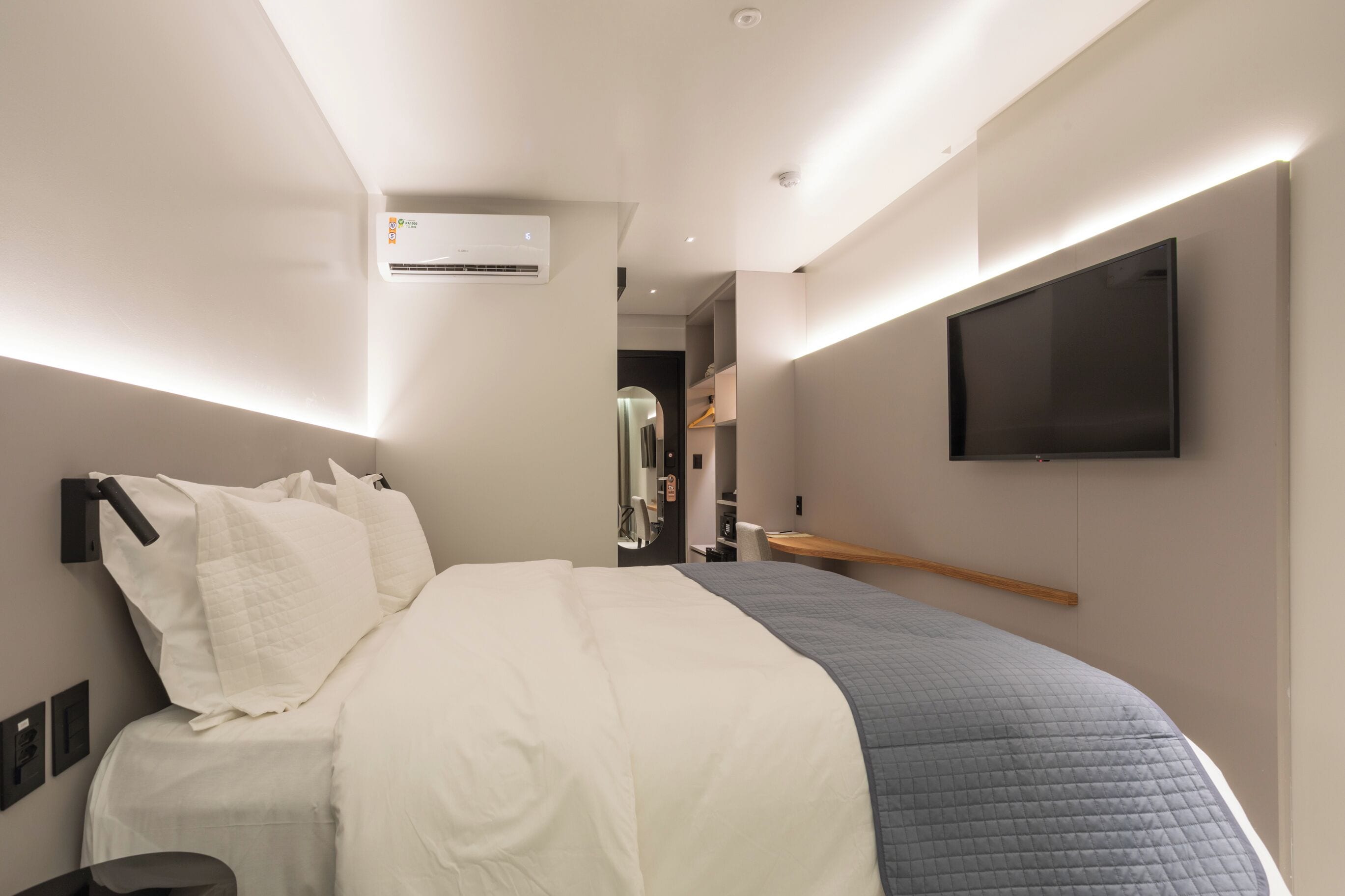 A modern hotel room with a large bed, a flat-screen TV mounted on the wall, and an air conditioning unit.