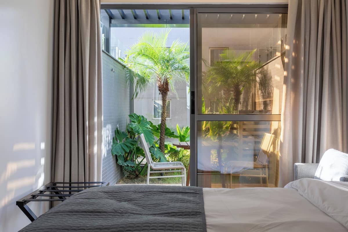 A bedroom with a bed, a chair, and a view of a balcony with plants and a palm tree.