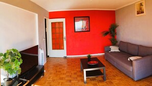 Een woonkamer met een rode muur, een grijze bank, een houten vloer, een televisie en een plant.
