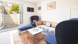 A living room with a sofa, a chair, a coffee table, and a TV. There is a door leading to an outdoor area with steps and a tree. - Host Stay Prince Albert (Margate)
