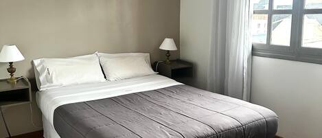 A bedroom with a large bed, two bedside tables with lamps, a window with curtains, and a ceiling light.