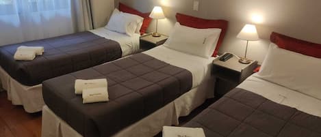 Ein Hotelzimmer mit drei Betten, jedes mit Kopfkissen und Decke. Nachttische mit Lampen und ein Fenster mit Vorhängen.