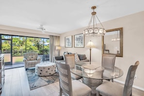 A modern living room with a glass dining table, comfortable seating, and a large window offering a view of greenery. - G-143 - First Floor With Lake Views -Sleeps 6 (Naples)