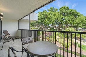 A balcony with a round table and two chairs, overlooking a garden with trees and a pathway. - E-233 - Deluxe Condo with Lake Views & Sleeps 6 (Naples)