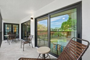A balcony with a table and chairs, large sliding glass doors, and a view of a garden. - E-233 - Deluxe Condo with Lake Views & Sleeps 6 (Naples)
