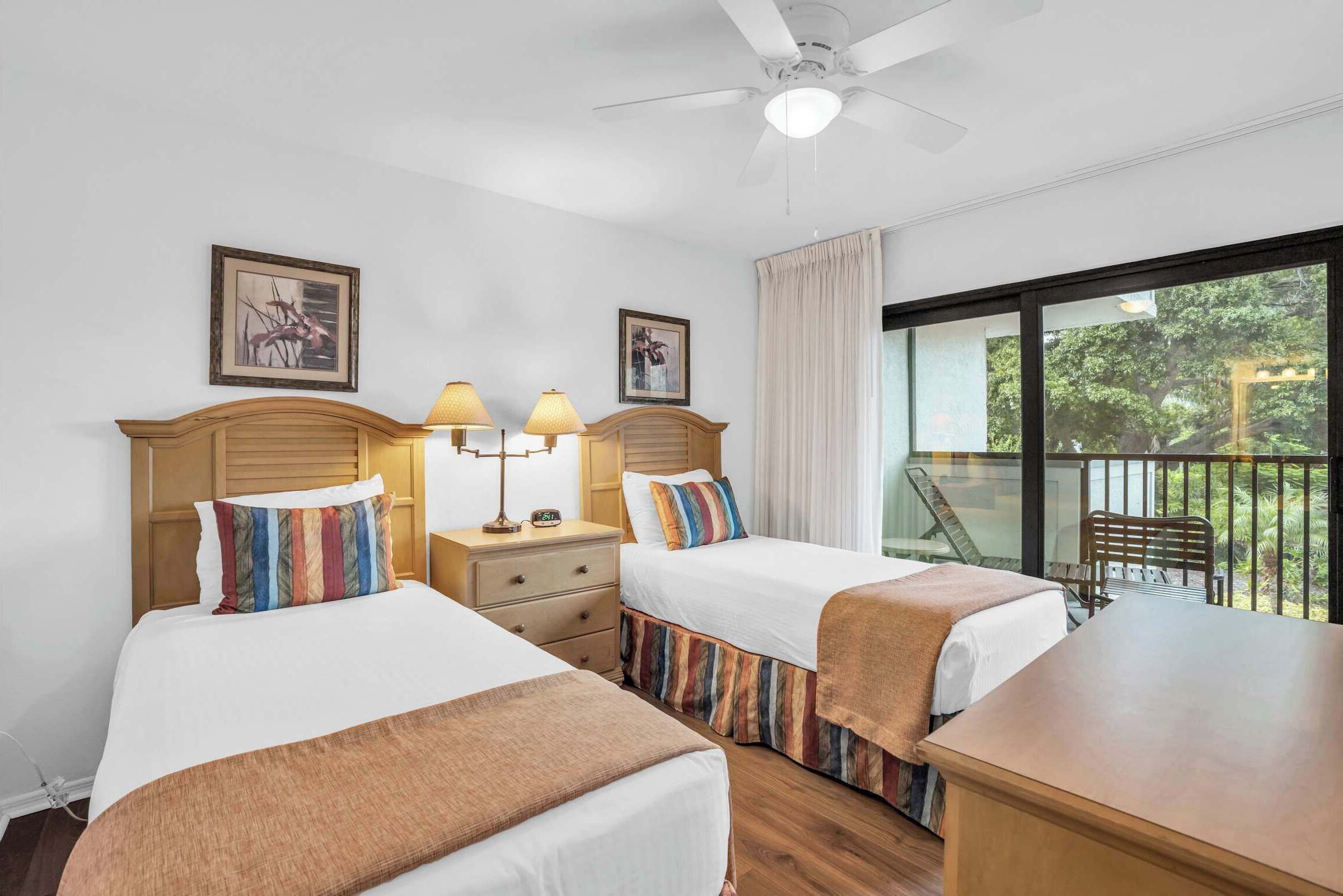 Habitación de hotel con dos camas, ventilador de techo y balcón con vista a zonas verdes.