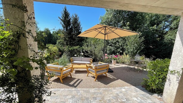 An outdoor patio with a wooden table and chairs, a beige umbrella, and a gravel floor. - Apartment LA BASTIDE (Rosières, Haute-Loire)