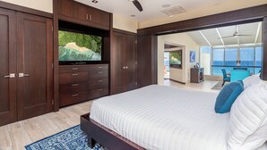 A bedroom with a large bed, a ceiling fan, a TV on a wooden cabinet, and a view of the ocean through a sliding door. - Puu Poa 407: 2  BR, 2  BA Condominium in Princeville, Sleeps 4 (Princeville)