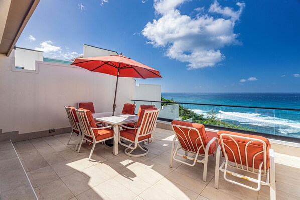 A balcony with a table and chairs, a red umbrella, and a view of the ocean. - Puu Poa 407: 2 BR, 2 BA Condominium in Princeville, Sleeps 4 (Princeville)