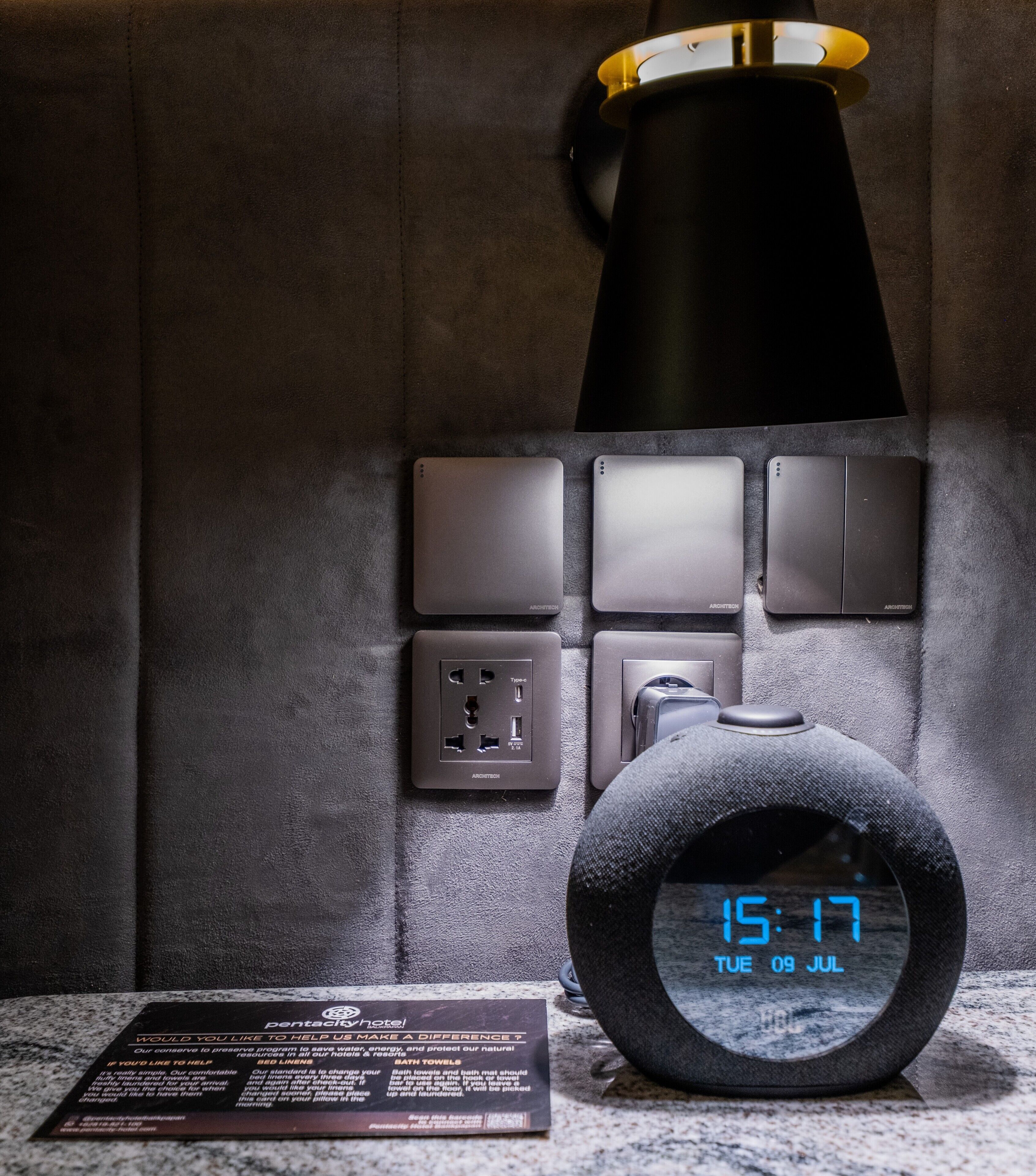 Een digitale klok waarop 15:17 staat, 9 juli, een zwarte lamp en een bord met informatie over de inspanningen van het hotel op het gebied van duurzaamheid.