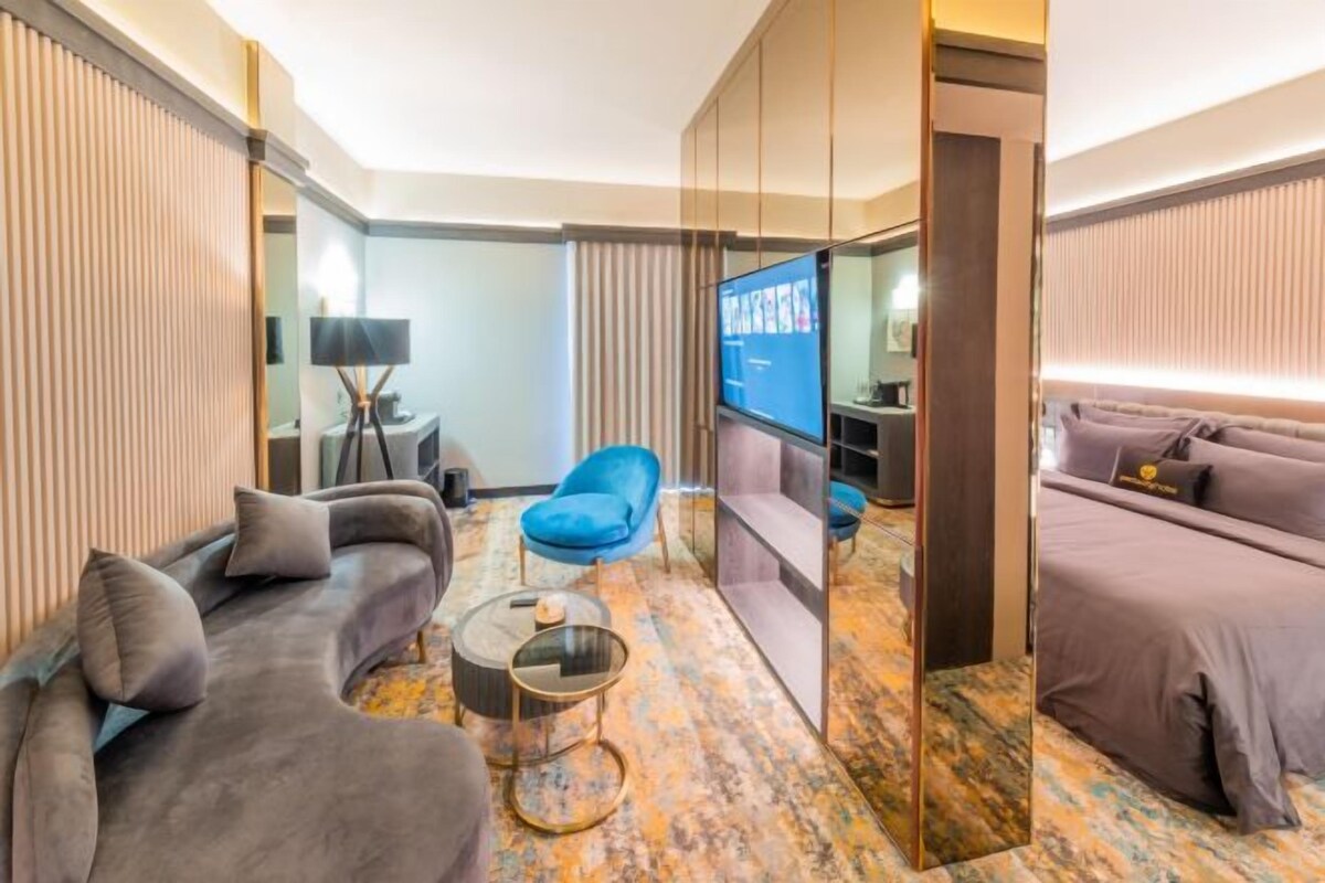 A modern hotel room with a sofa, a blue armchair, a TV, and a bed.