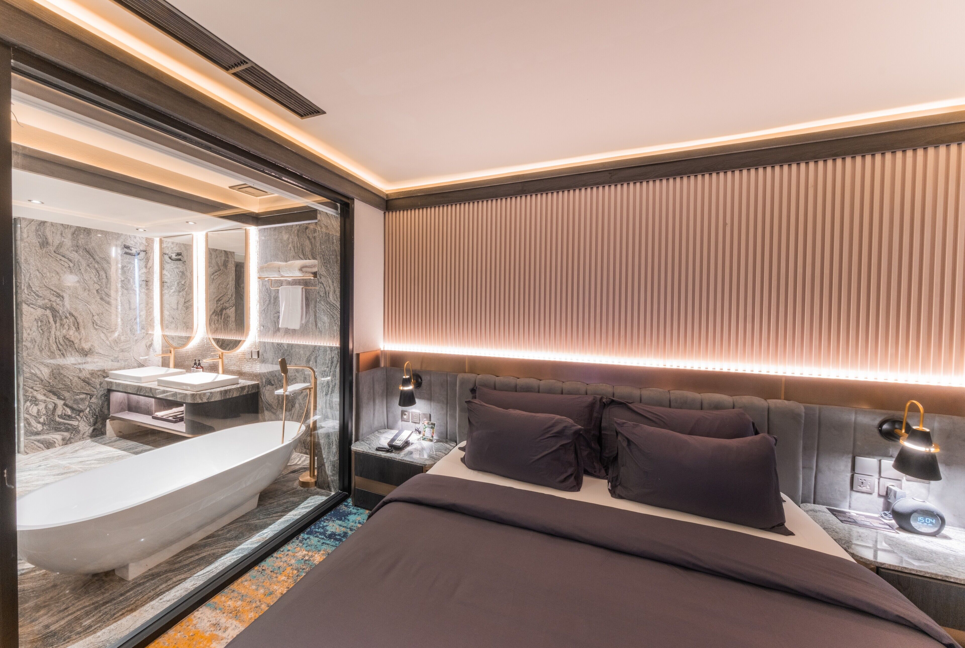 A modern hotel room with a large bathtub, a bed with dark bedding, a bedside table with a lamp, and a wall-mounted mirror.