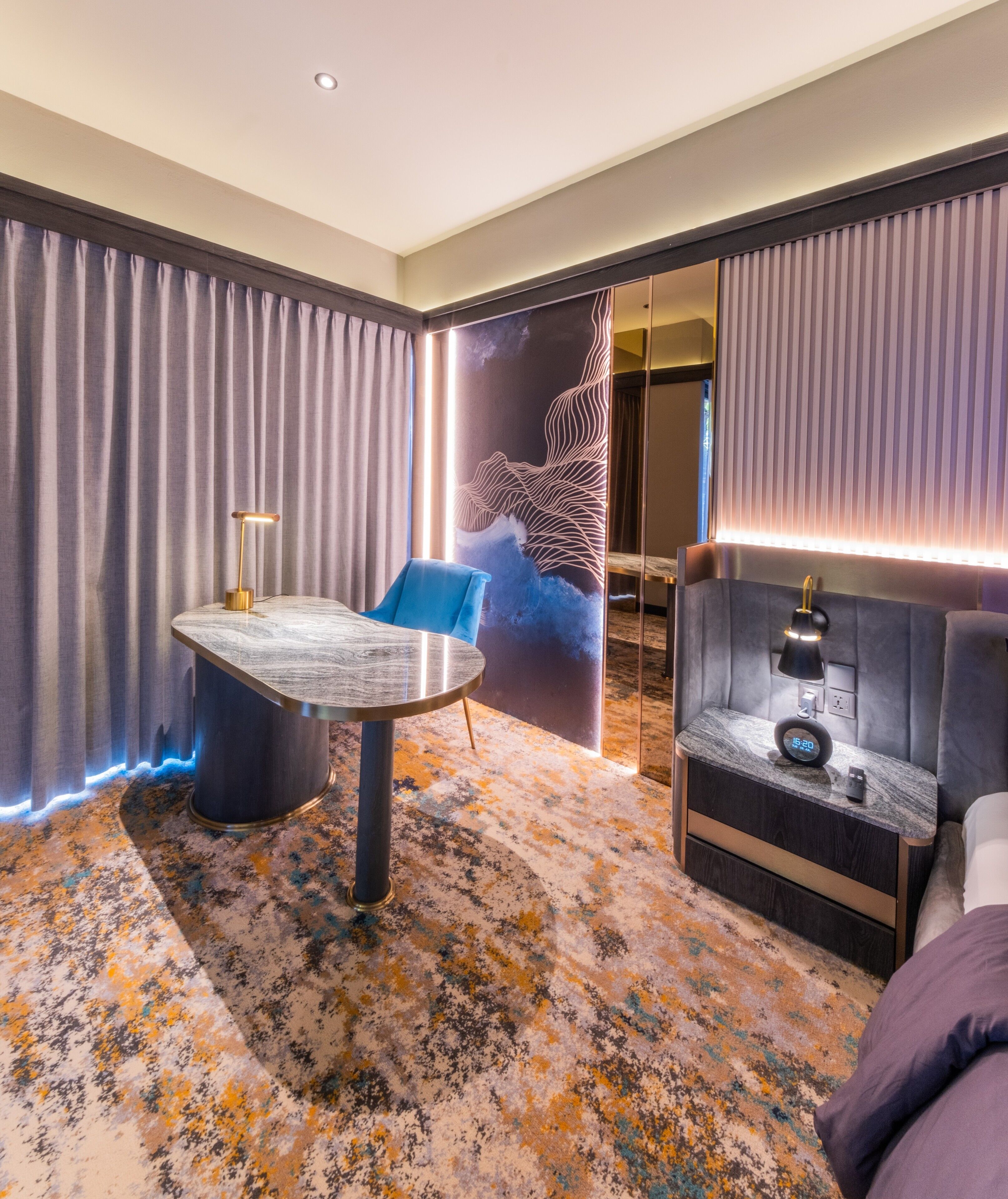 A modern hotel room with a round table, a blue chair, a bed, a nightstand with a clock, and a decorative wall mural.