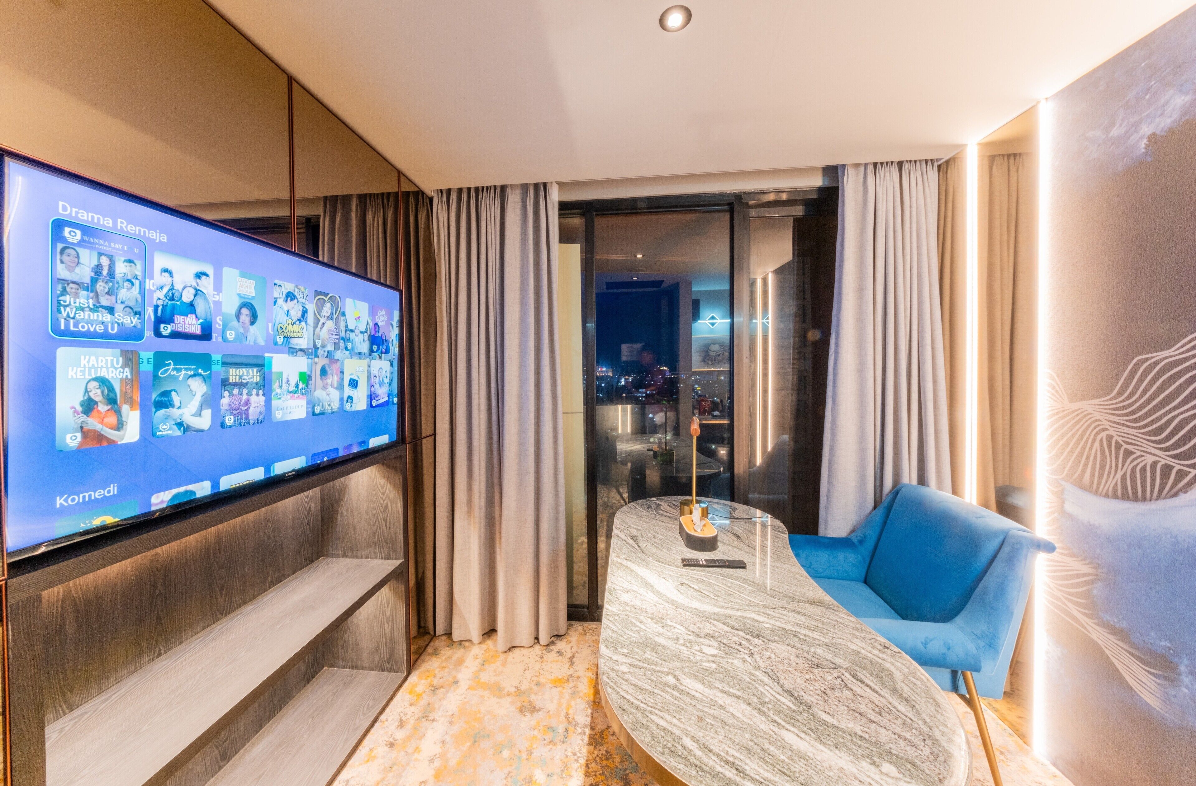 Habitación de hotel moderna con una televisión grande mostrando opciones de películas, un sofá azul, una mesa de mármol y vistas de la ciudad a través de la puerta de cristal.