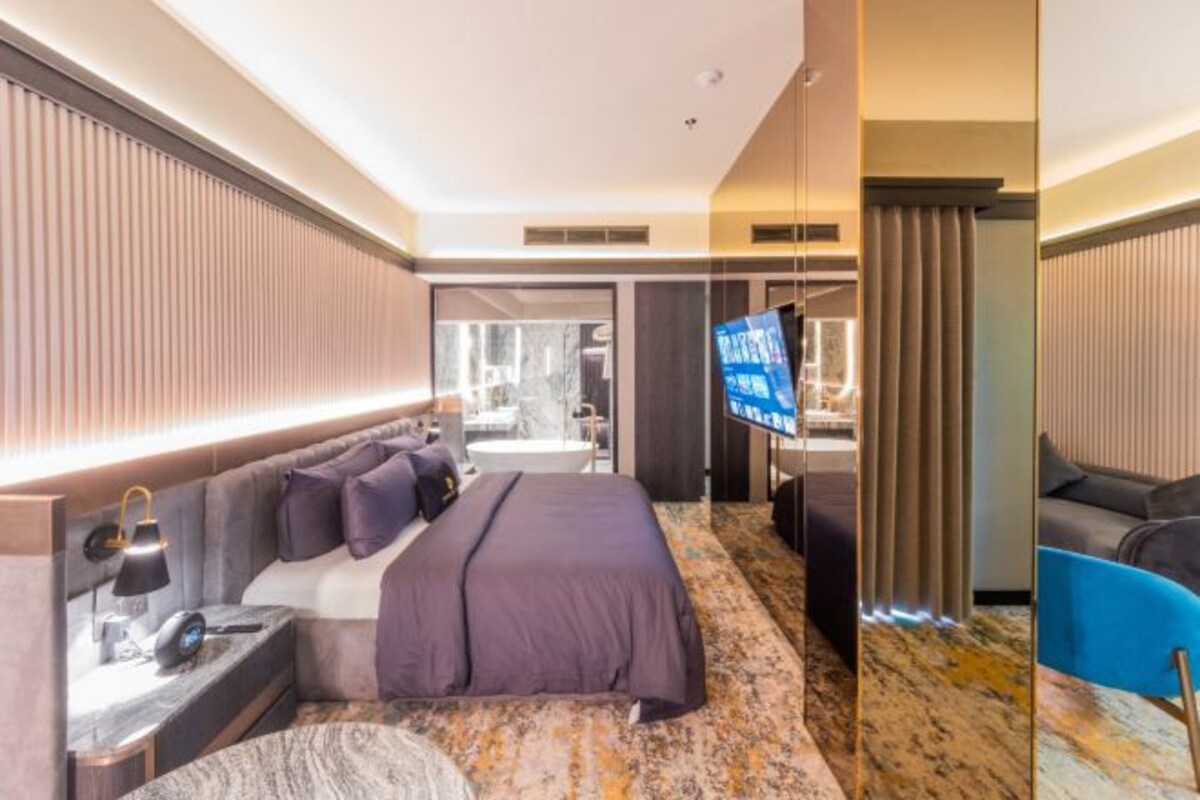 Quarto de hotel moderno com uma cama grande, televisão, escrivaninha e banheiro visível por uma porta aberta.
