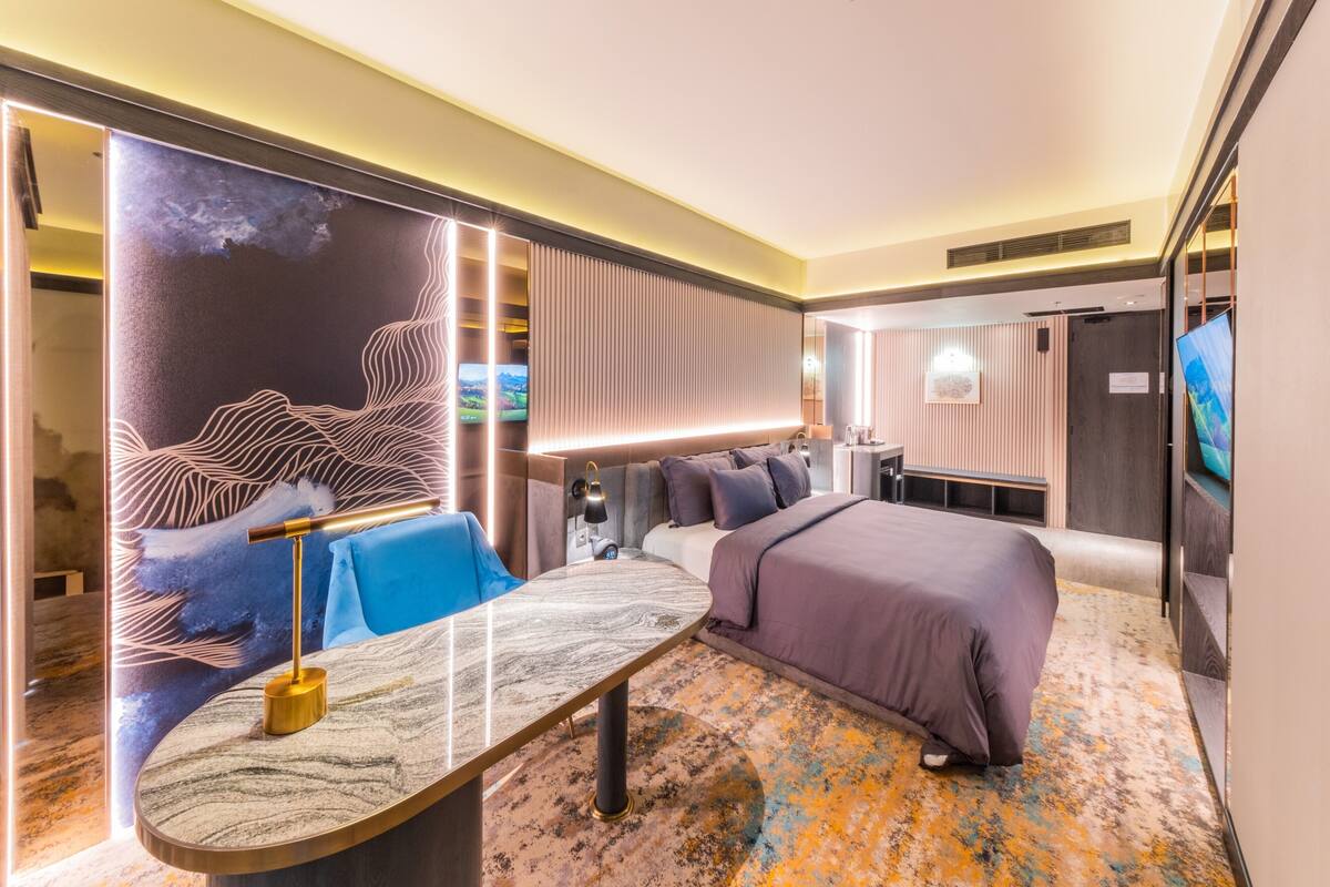Een moderne hotelkamer met een groot bed, een bureau met lamp, een zithoek met een blauwe bank en een groot kunstwerk aan de muur.
