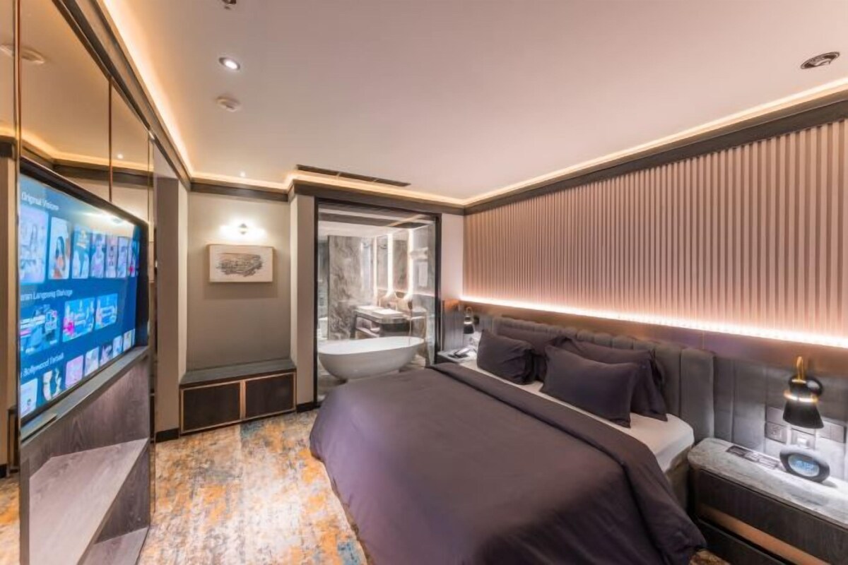 A modern hotel room with a large bed, a flat-screen TV, a bathroom with a bathtub, and a wall-mounted light fixture.