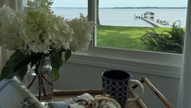 A tray with a plate of cinnamon rolls, a cup of coffee, and a newspaper by a window with a view of a body of water and a dock.