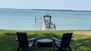 A lakeside view with a wooden dock leading to a gazebo, two Adirondack chairs, and a round table on a grassy area.