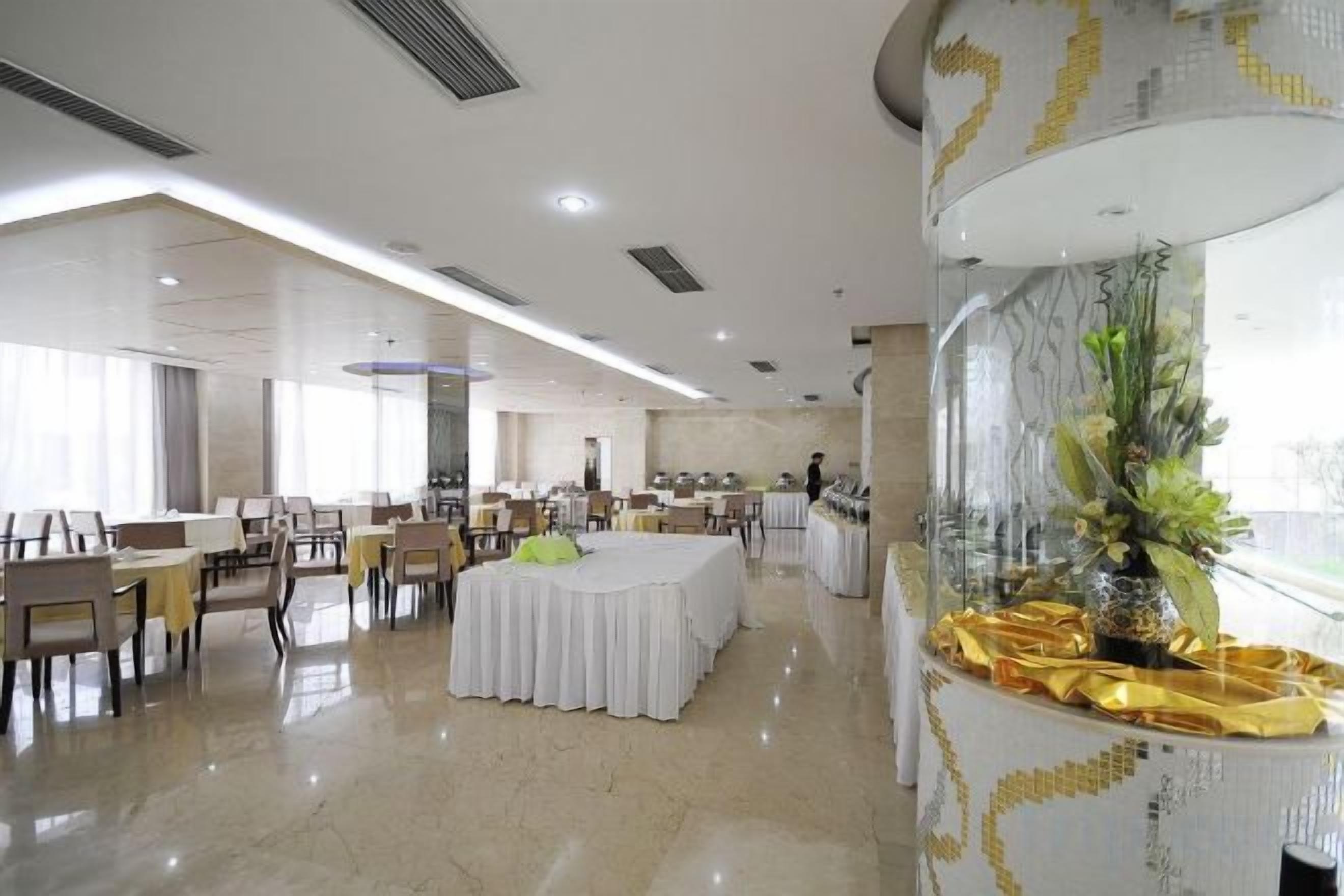 A spacious hotel dining area with round tables and chairs, a central decorative plant, and a large window providing natural light.