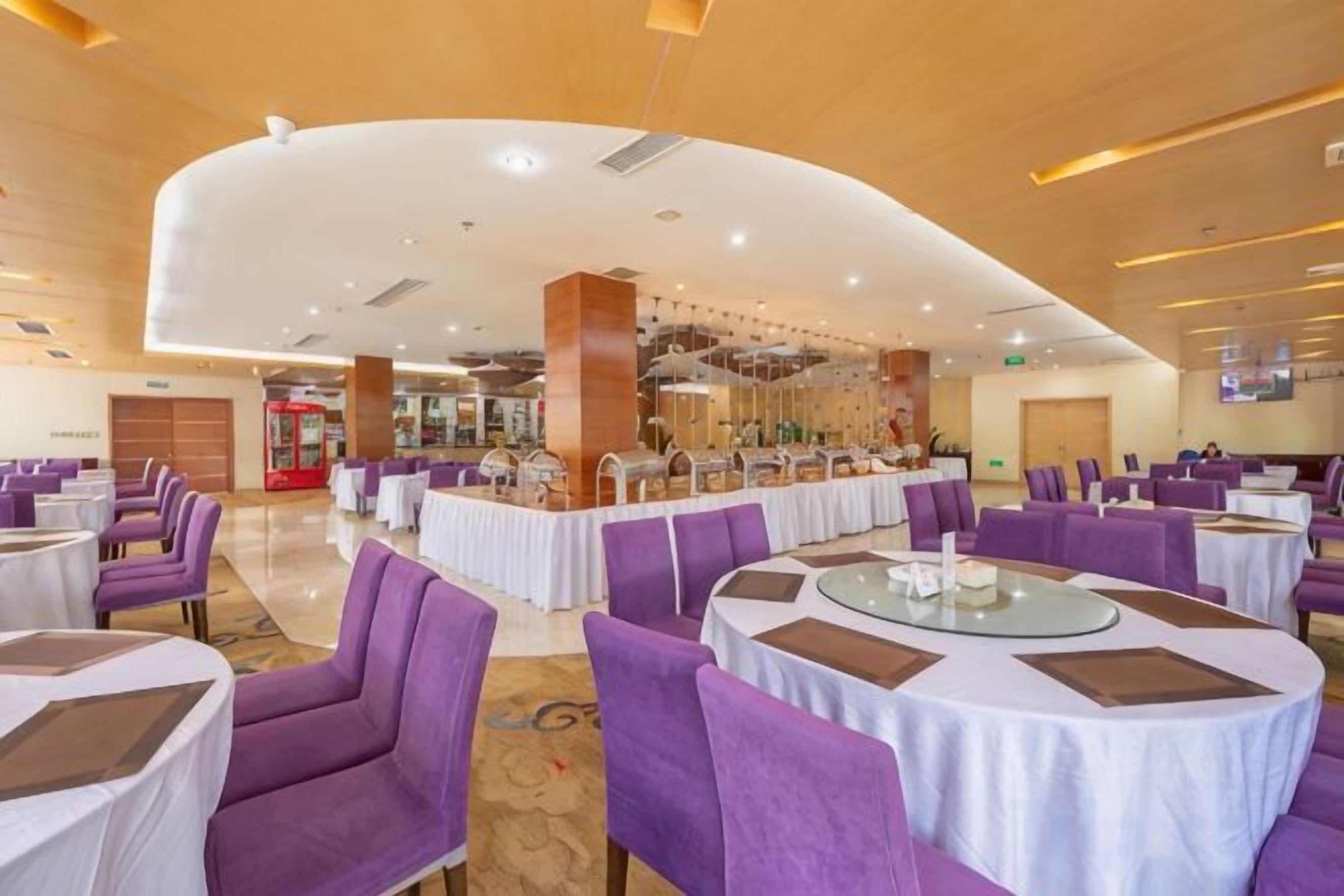 A hotel dining area with round tables and purple chairs, a central bar area, and a modern ceiling design.