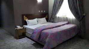 A hotel room with a bed, a nightstand, and a window with curtains. - Silicon Valley Resort (Enugu)