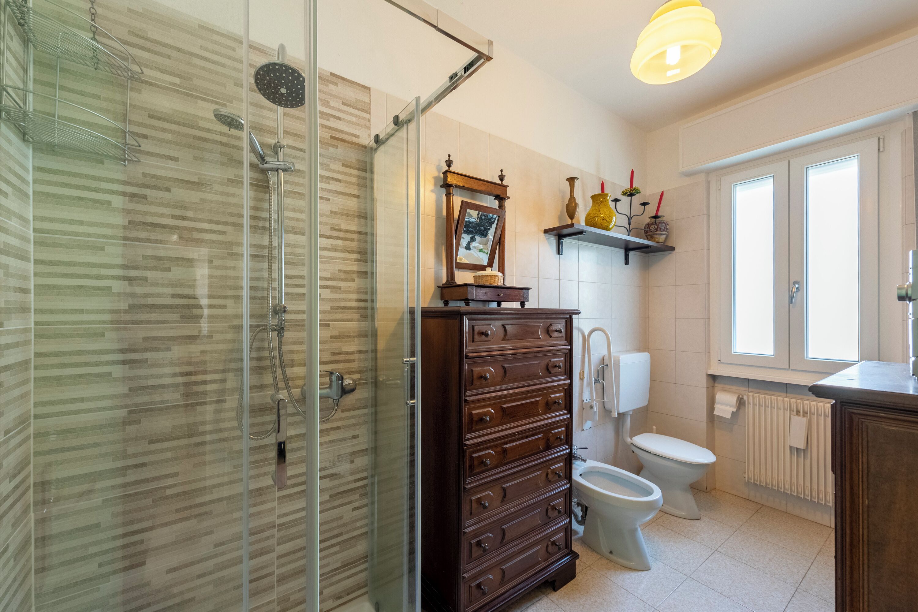 Une salle de bain dotée d’une douche à parois de verre, d’une commode en bois, de toilettes et d’une fenêtre laissant entrer la lumière naturelle.