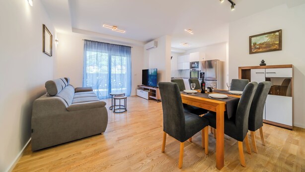 Een moderne woonkamer met een bank, eettafel en stoelen, een keuken op de achtergrond en een groot raam met gordijnen.