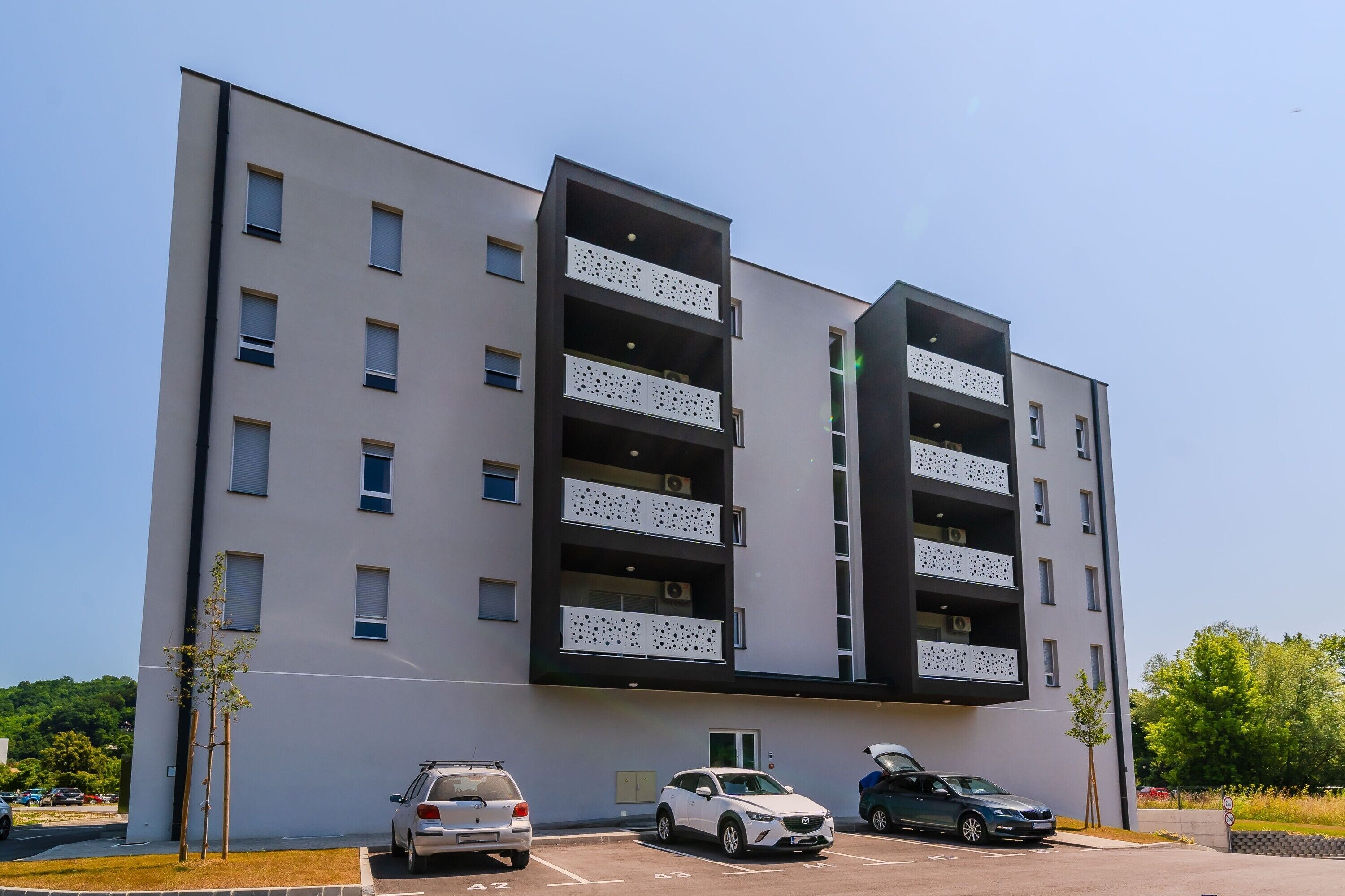 A modern multi-story building with balconies, a parking area, and a clear sky.