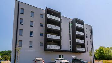A modern multi-story building with balconies, a parking area, and a clear sky.