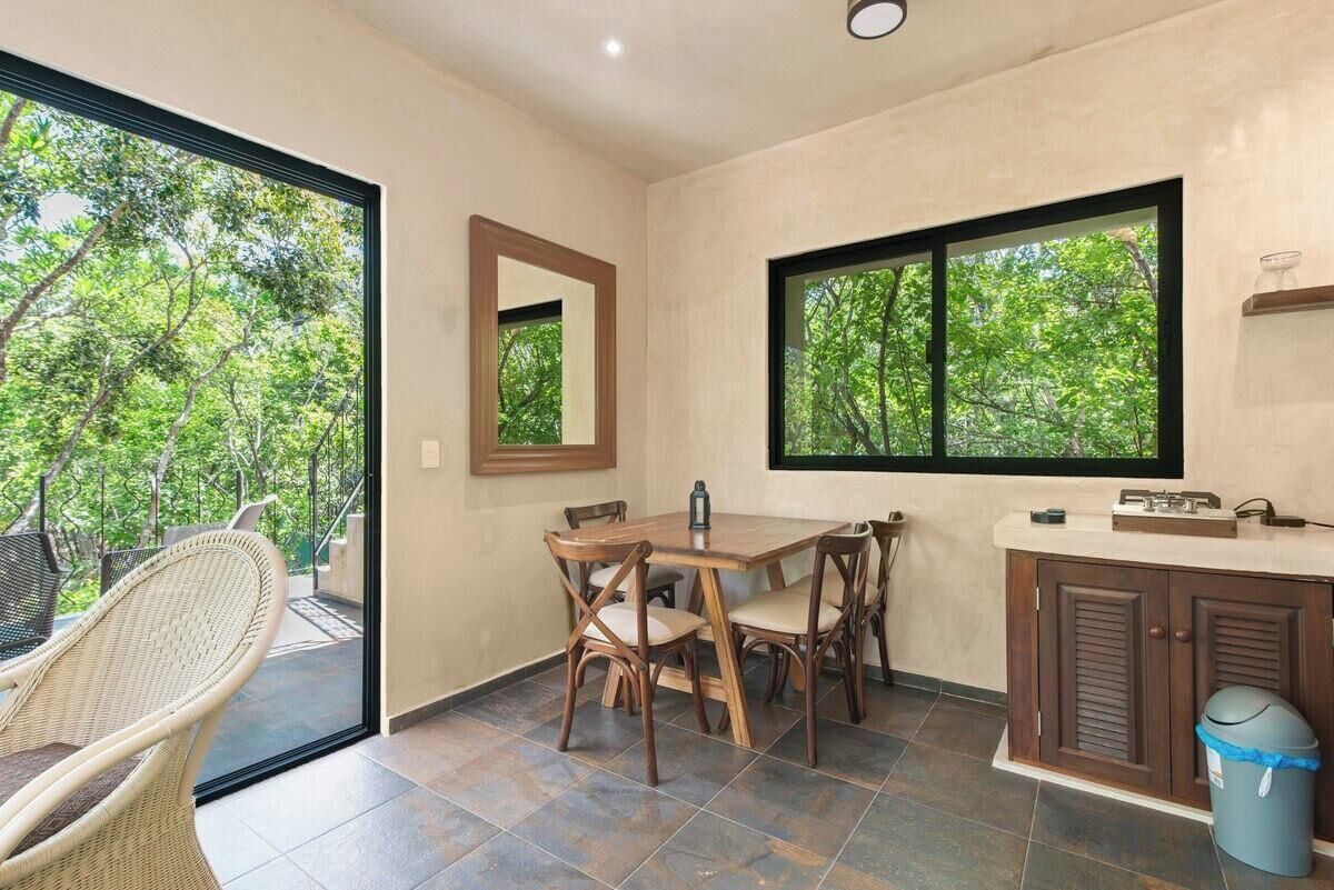 Un coin repas avec une table et des chaises, une fenêtre donnant sur des arbres et un meuble contenant une poubelle.