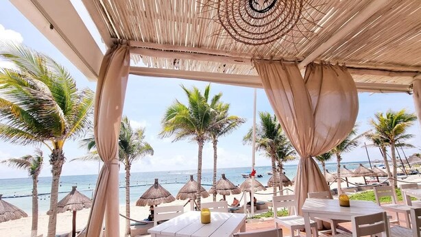Un espace extérieur aménagé avec des tables et des chaises blanches, offrant une vue sur une plage bordée de palmiers et dotée de parasols en chaume.