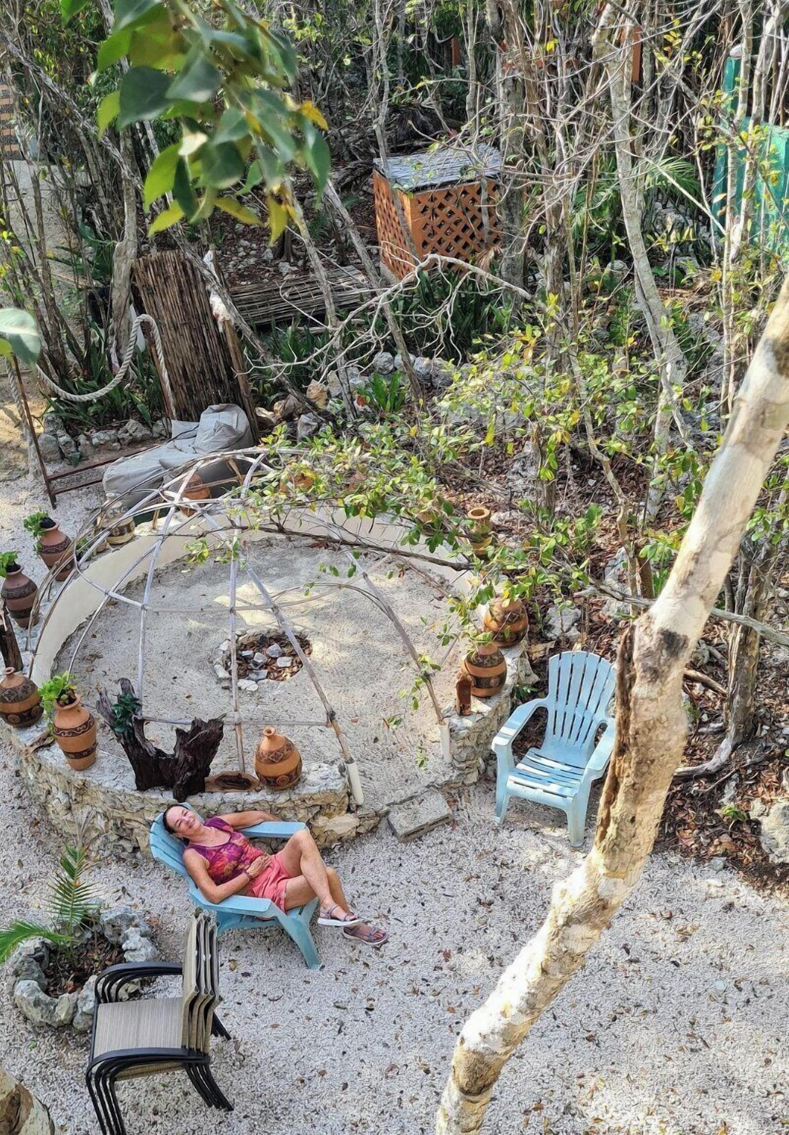 Ein Gartenbereich mit Schaukelstuhl, blauem Stuhl und einer Hängematte. Im Hintergrund sind Blumentöpfe und ein kleines Gebäude zu sehen.