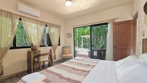 Une chambre avec un grand lit, un bureau et une chaise. Il y a une fenêtre avec des rideaux, ainsi qu’une porte donnant sur un espace extérieur.