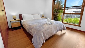 A bedroom with a large bed, a nightstand, a bedside table, and a window with a view of greenery.