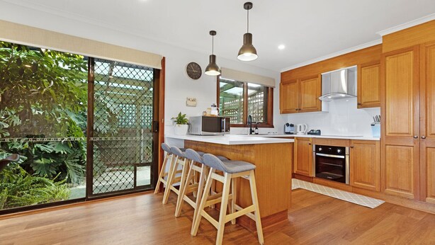 A modern kitchen with wooden cabinets, a central island with bar stools, a large window, and a view of greenery outside.