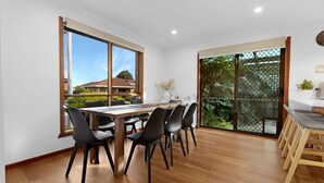 A dining area with a wooden table and black chairs, large windows with a view of houses, and a sliding glass door leading to a garden.