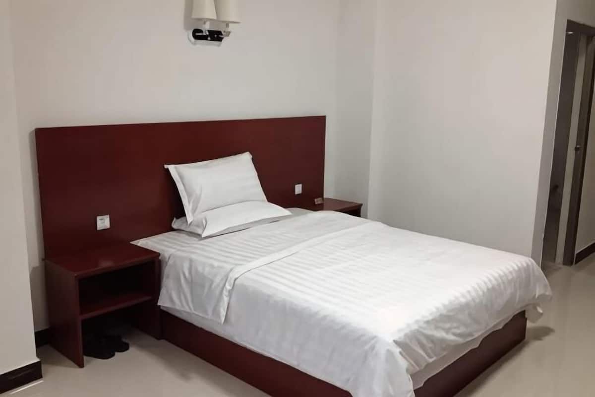 Una cama individual con cabecera de madera, una colcha blanca y una mesita de noche con una lámpara.