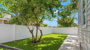 Un patio trasero cercado con césped sintético, un árbol y un camino pavimentado.
