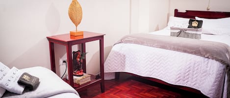 Habitación de hotel con piso de madera, una mesita de noche con un objeto decorativo, una cama con colcha acolchada y una lámpara fijada a la pared.
