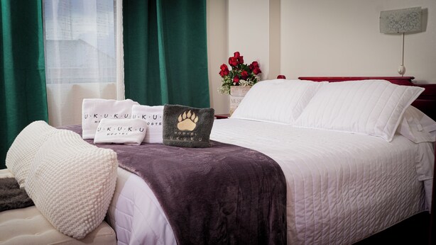Una cama bien hecha con ropa de cama blanca, un edredón morado y una almohada con diseño de huella. Hay toallas blancas con el texto "U.K.U.K.U HOSTEL" y un jarrón con rosas rojas sobre la mesita de noche.