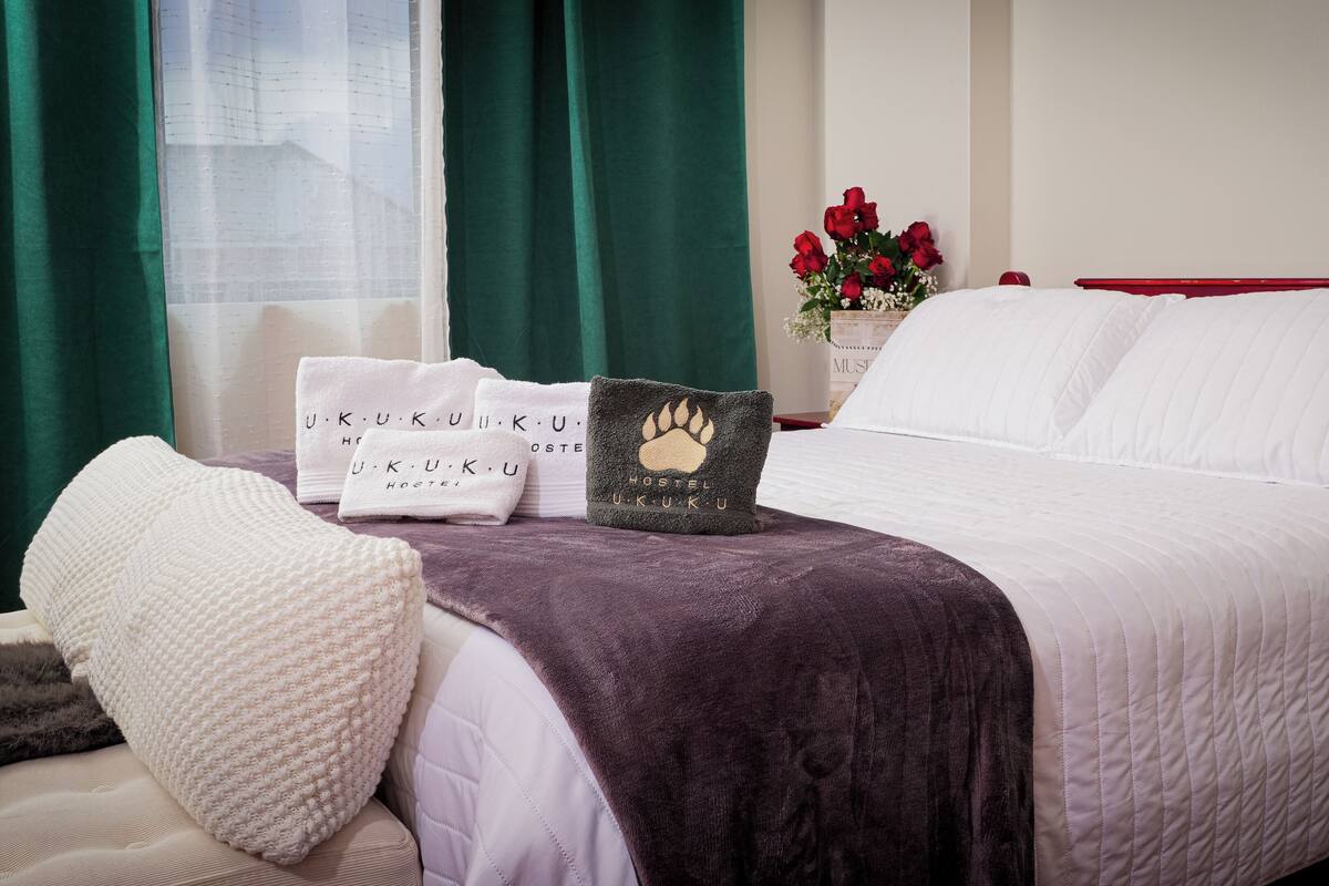 A neatly made bed with white linens, a purple throw blanket, and a pillow with a paw print design. There are white towels with the text "U.K.U.K.U HOSTEL" and a vase with red roses on the nightstand.