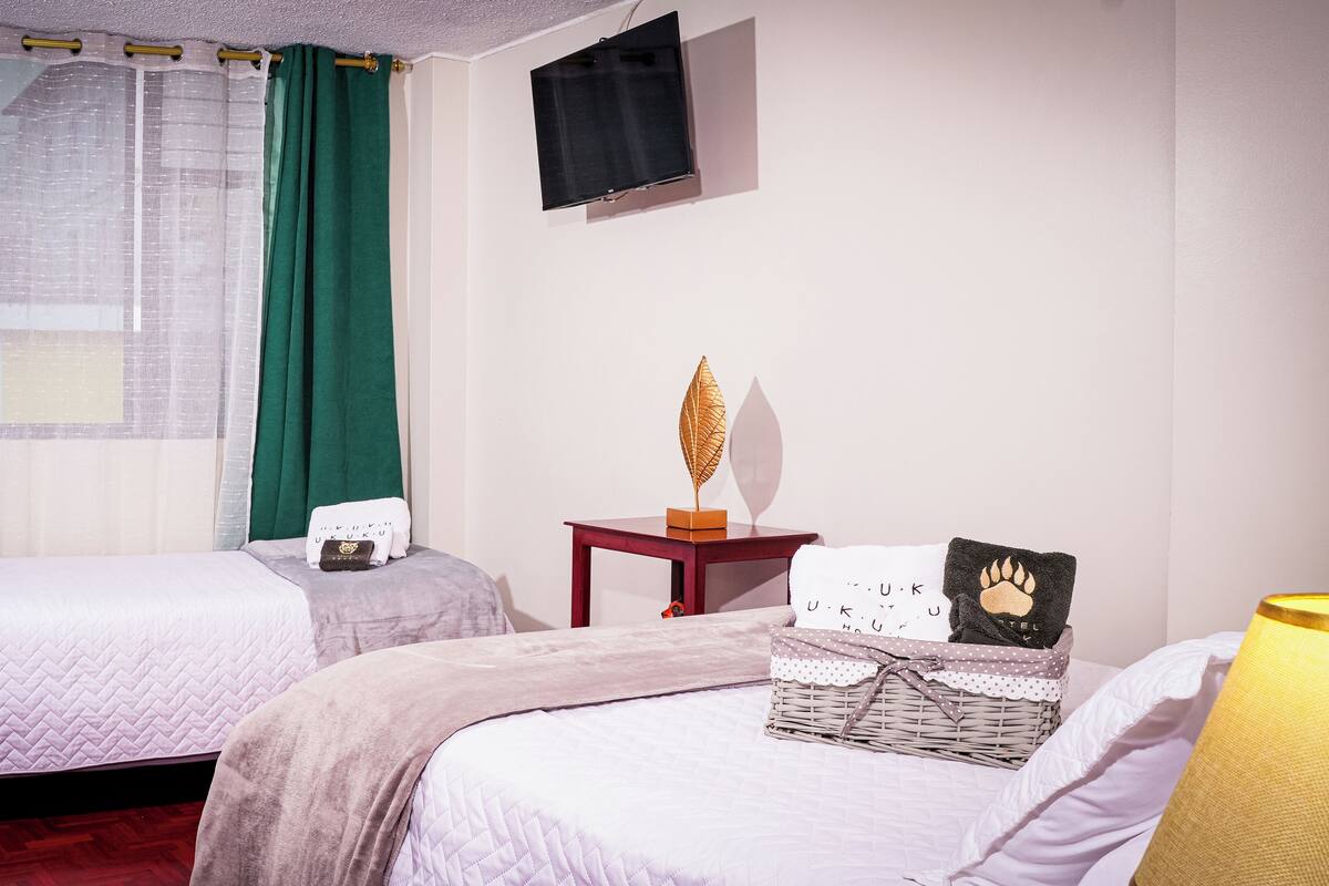 A hotel room with two beds, a small table with a decorative item, a wall-mounted TV, and a window with green curtains.