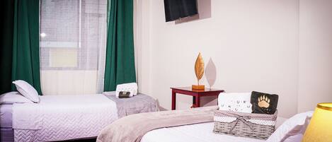 A hotel room with two beds, a small table with a decorative item, a wall-mounted TV, and a window with green curtains.