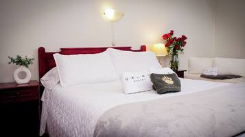 Una cama bien tendida con ropa de cama blanca, cabecera roja y una mesita de noche con un jarrón de flores y una lámpara.