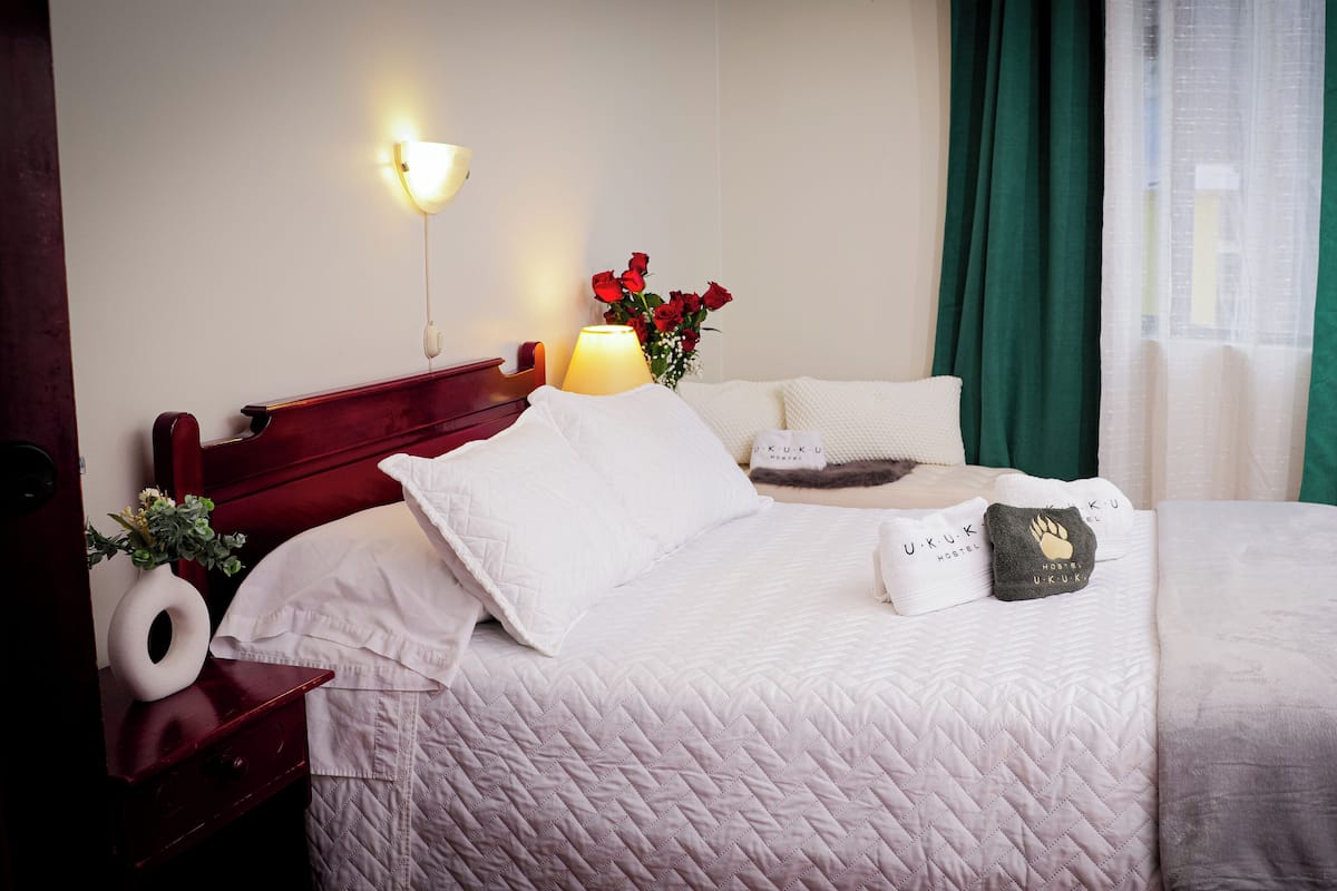 A neatly made bed with white bedding, a bedside lamp, a vase with red roses, and a wooden nightstand with a white towel and a white vase with green foliage.