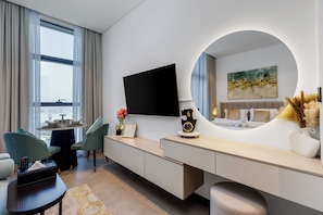 A modern hotel room with a large window, a flat-screen TV, a round mirror, a desk, and a bed with a headboard.
